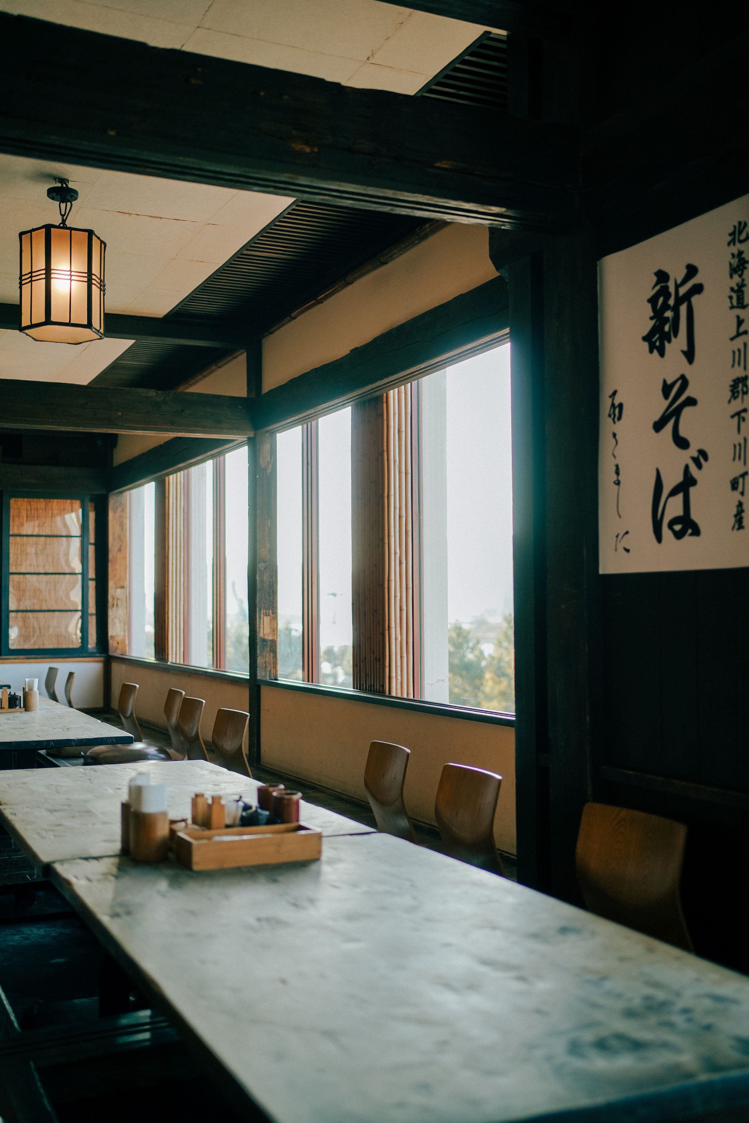 窓から自然光が差し込む、木のテーブルが並ぶ飲食店の空間